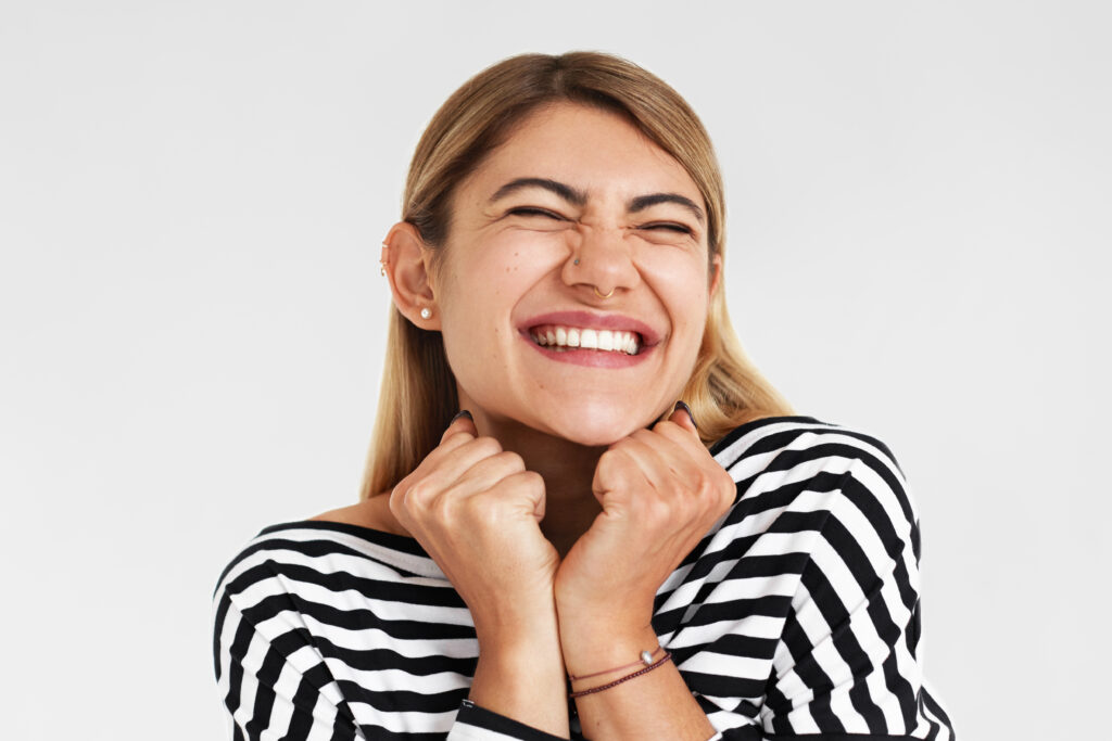 portrait of beautiful emotional overjoyed young woman wearing striped top beaming with happiness and screwing up her eyes, holding clenched fists at face, excited with success, winning or victory
